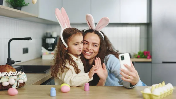 A cheerful family is preparing for the easter holiday using smartphone to video calling. Mom and daughter paint eggs, communicate with friends through video. Online talking. Happy easter