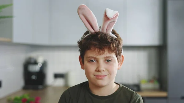 Portrait of the smiling little boy wearing bunny ears headband sitting in the kitchen table looking at the camera. Happy easter. Child celebrate Easter holiday