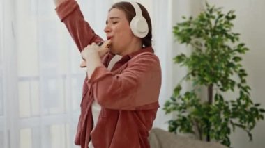 Happy Young Woman in Headphones Dancing and Singing in Smartphone Having Fun in Living Room. Girl Singing and Looking at Camera. People, Lifestyle and Joy Concept