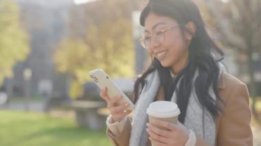 Smiled Asian Woman Walking in the Park with Cup of Coffee Using Smartphone. Beautiful Girl Chatting with Mobile Outside. Female Traveler. People and Technology Concept