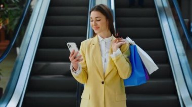 Caucasian Girl Standing in Shopping Mall, Using App Smartphone and Holding the Paper Bags. Female Customer After Shopping Texting Chatting on Mobile Phone. Happy Woman Purchase