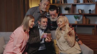 Group of mature people enjoy using smartphone and sitting on sofa in nursing home. Elderly man showing something to his friends, scrolling apps. Senior people spending time together
