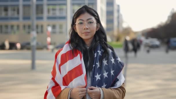 骄傲的微笑站在街头的亚洲女人被美国国旗包裹看看相机 人民支持美国的民主 外面的爱国女士肖像女孩庆祝国庆节 特写镜头 — 图库视频影像