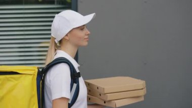 Teslimat servisi. Beyaz şapkalı, sarı sırt çantası taşıyan, üzerinde üniforması olan, elinde pizza tutan Kafkasyalı Kurye Kız 'ın yan görüntüsü. Fast Food Teslimatı.