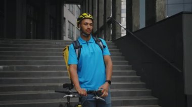 Yakışıklı Afro-Amerikan Kurye portresi Ofis Merkezi 'nin yanında bisikletiyle duruyor, Kameraya bakıyor ve Sarı Sırt çantalı Üniforma giyiyor. Teslimat Hizmeti