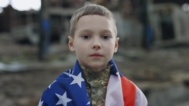 身穿军服的小爱国战士站在废墟背景下的美国国旗上 男基德拿着美国国旗看着摄像机 爱国者 特写镜头 — 图库视频影像