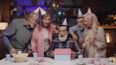 Doğum günü şapkalı heyecanlı yaşlı erkek, hediyeyi açıyor, huzurevindeki diğer yaşlılarla birlikte kutluyor. Doğum günü partisi, hediyeler, Eski Dostlar Üfleme Partisi Kornaları