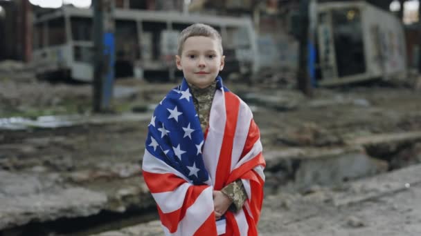 身着军服的小爱国男兵站在美国国旗上 在废墟的背景下凝视着相机 肖像画小子爱国者 — 图库视频影像
