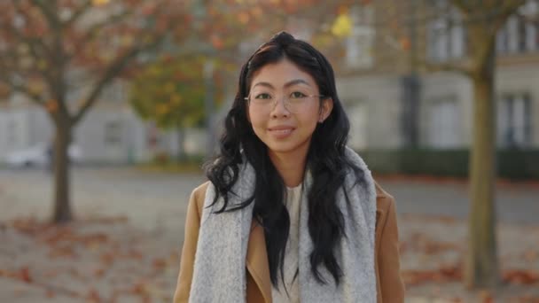 眼鏡をかけたアジア系の魅力的な少女の肖像画 人の顔 女性カメラを見て秋の街の背景に立っている 外の若いビジネスウーマン — ストック動画