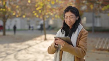 Kulaklık takan gülümseyen Asyalı genç kadın, Şehir Tableti kullanarak açık havada duruyor. Sokakta çalışan şık bayan, kaydırma uygulamaları. Çevrimiçi Kız İzleme Alanı. Teknoloji ve İş Konsepti