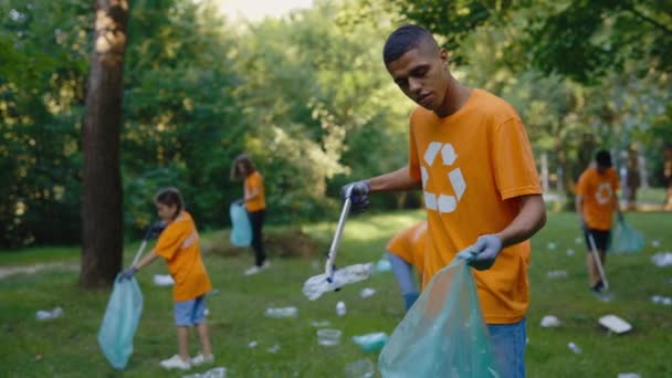 多様なボランティアのグループ公共公園の清掃を象徴するリサイクルTシャツ ごみ箱袋にゴミを収集しながら ゴミ箱ピッカーを使用して多民族の男活動家 安全な生態学的概念 — ストック動画