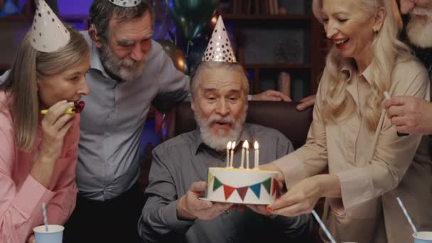 戴着生日礼帽的老人在生日蛋糕上吹灭蜡烛 和其他老人一起在养老院庆祝 生日聚会 在一起消磨时光 吹喇叭 — 图库视频影像