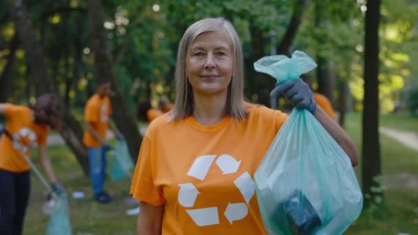 白种人老年女性生态义工穿着T恤衫 提着装满垃圾的垃圾袋站在公园里的肖像 生态活跃分子看着相机 安全生态概念 — 图库视频影像