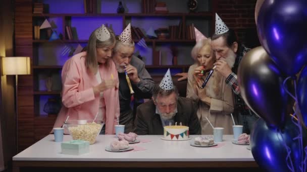 在疗养院的餐桌上 坐在宴会桌旁的老年人 和朋友们一起庆祝生日时 在蛋糕上放蜡烛 长者在一起消磨时间 吹喇叭 — 图库视频影像