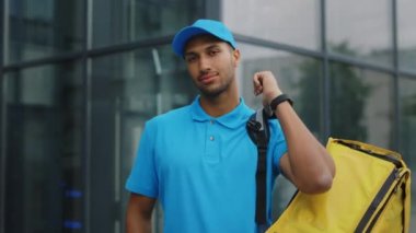 Ofis binasının yanında duran sarı sırt çantasını taşıyan mavi üniformalı Afro-Amerikan erkek kurye gülümsedi. Kameraya bakan çok ırklı bir postacı. Halk ve Meslekler. Teslimat Hizmeti