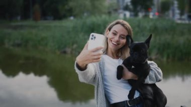 Güzel Mutlu Kadın, sevimli siyah buldoguyla gölün yanında duruyor ve köpeğiyle selfie çekiyor, köpeğiyle anılar yaratıyor. Teknoloji ve İnsanlar Dostluk Konsepti