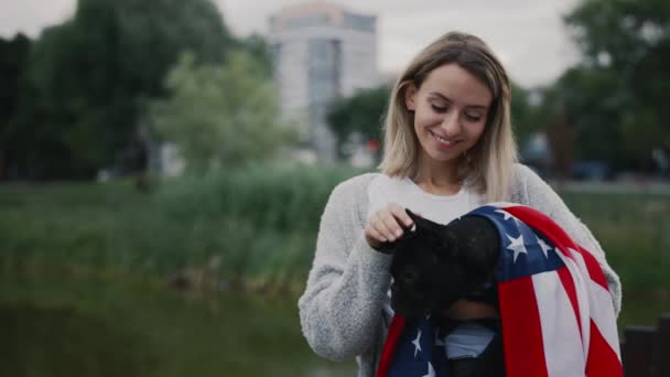 在公园外 白人女店主牵着她的黑色小斗牛犬微笑着 把美国国旗挂在外面 人与狗的爱国主义概念 — 图库视频影像