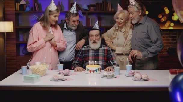誕生日の帽子のシニア男性は 退職家で他の高齢者と一緒にお祝いの誕生日ケーキにキャンドルを吹いています 誕生日パーティー 一緒に過ごす時間 — ストック動画