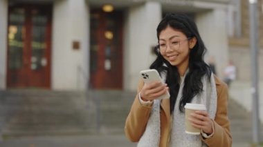 Gülümseyen Asyalı Kız dışarıda akıllı telefon kullanıyor ve kahve içiyor. Kahve molası. Cep Telefonu Uygulaması ile İnternet 'te Sörf Yapan Güzel İş Kadınları.