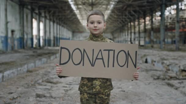 破壊された工場に立って寄付サインを保持している若い子供兵士の肖像画 子供は助けを求める カメラを見て難民はほとんどいない 戦争問題 — ストック動画
