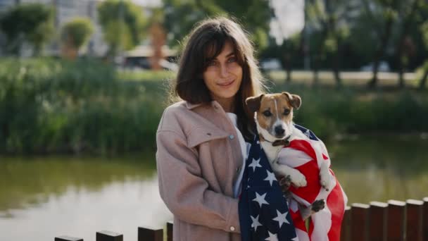 在美国国旗上挂着她的小杰克罗素的白人女房东面带微笑的画像 站在公园外面看相机的女孩 人与狗的爱国主义概念 — 图库视频影像
