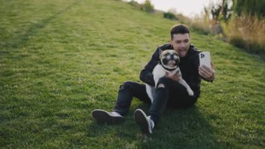 Yakışıklı, gülümseyen beyaz adam Brooklyn bulldoguyla çimenlerde oturuyor ve köpeği gibi selfie çekiyor, evcil hayvanıyla anılar yaratıyor.