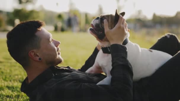 快乐的白人主人躺在公园里躺着 和他的布鲁克林斗牛犬玩耍 玩得开心 男人抚摸他的宠物 表达爱 人与狗的友谊概念 — 图库视频影像