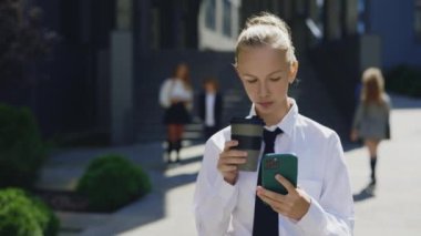 Genç bayan küçük patron ofis binasının yanında yürüyor, akıllı telefon kullanıyor, kahve içiyor. Çevrimiçi Arkadaşlarla Çocuk İş Kadınları Ağı Uygulamaları Kapıyor, Dışarıya Mesajlar Yazıyor