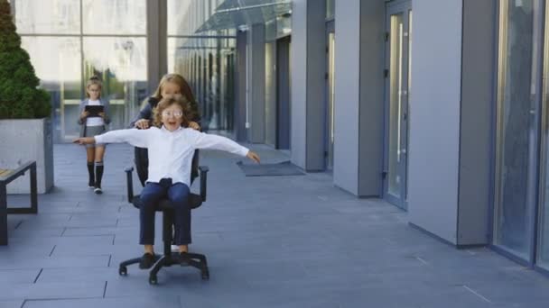 近くのオフィスビルの外のオフィスチェアで楽しい乗馬をしている興奮した小さなビジネスマン ビジネスキッズが休憩し ゲームを楽しみ 現代のビジネスセンターで一緒に笑う — ストック動画