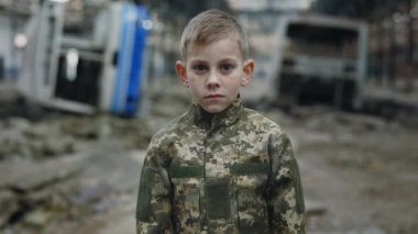 Askeri üniforma giyen küçük asker yıkıcı arka planda dikiliyor ve kameraya bakıyor. Yok Edilmiş Bitki 'de Erkek Mültecilerin Çocuk Portresi. Ukrayna Savaşçısı. Savaş