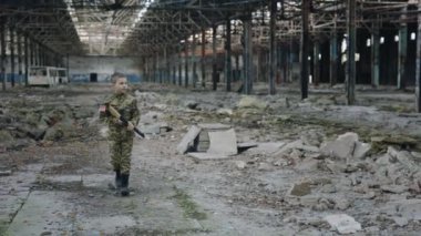 Küçük Amerikan askeri harabelerin arkasında oyuncak silah tutan tahrip edilmiş bir tesiste yürüyor. Görevdeki Savaşçı. Askeri giysi. - Savaş. Amerikan vatanseveri. Modern Savaş ve Savaş Konsepti