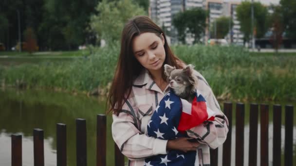 照片上 这位高加索女人的主人微笑着 手里拿着裹着美国国旗的吉娃娃 站在公园外看着摄像机 人与狗的爱国主义概念 — 图库视频影像