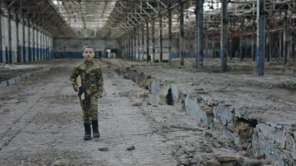 破壊された植物の中を歩く軍の制服の少年遺跡の背景におもちゃの銃を持っています パトリオット兵士キッド 戦争で遊ぶ少年 — ストック動画