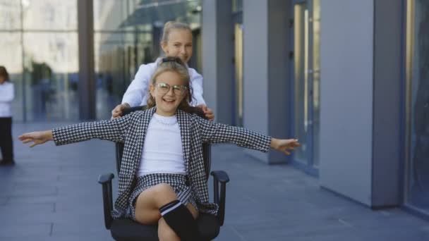 仕事の後に楽しい時間を過ごしている2人の幸せな小さなビジネスマン 小さな女の子はオフィスの椅子に座って 彼女の同僚は ビジネスセンターの近くに彼女を乗って 休憩中の生徒たち — ストック動画