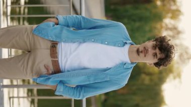 Dikey video. Beyaz Sakallı Yakışıklı Adam caddenin dışında dikiliyor ve kameraya bakıyor. Caddede duran keyifli erkek portresi. Doğa Gölü Arkaplanı