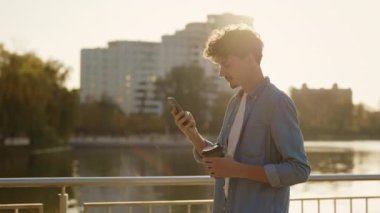 Gün batımında sokakta yürürken, akıllı telefonunu kullanarak arkadaşlarıyla mesajlaşırken yan görüntüsü. Kafkasyalı erkek Yürürken Gezici Kaydırma Aracı