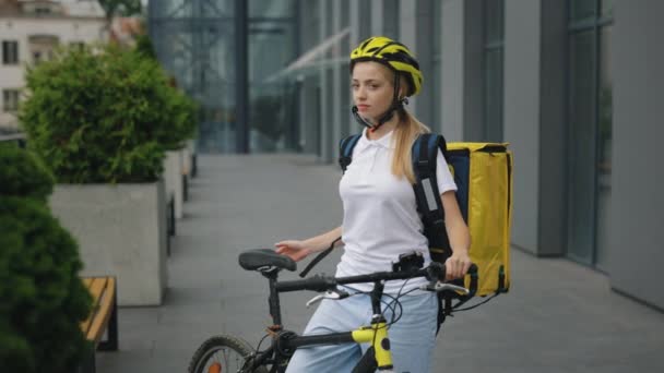 バイクの近くに黄色のバックパックとヘルメットが立っているかなり若い配信女性の肖像画 街中で食べ物や飲み物を届ける女性宅配便カメラを見る — ストック動画