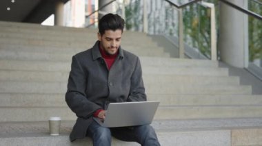 Yakışıklı Sakallı Çoklu Etnik Adam dışarıda merdivenlerde oturuyor, dizüstü bilgisayarla çalışıyor. İş adamı antrenman yapıyor. Yabancı Öğrenci Bilgisayar Kullanarak Sokakta Çalışıyor