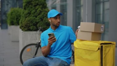 Yakışıklı, Afro-Amerikan Postacı, Mavi Üniforma Giyiyor ve Şapka Giyiyor, Dışarıda Oturuyor, Teslimat Paketinde Barkod Taranıyor. Man Courier tarayıcı kullanıyor. Yönetici