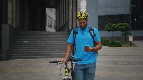 非裔美国人信使步行在办公大楼附近 使用智能手机 送货应用程序 穿着特别的蓝色制服 背着黄色背包和助手 单车派递 — 图库视频影像