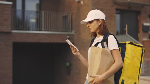 快递员手拿着纸包和手机沿着大楼走着 送货女士在街上寻找送货地址 食物送到户外时使用智能手机的可爱女人 — 图库视频影像