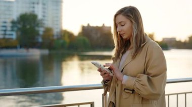 Gün batımında Sokakta Yürüyen Genç Kadın 'ın yandan görünüşü, akıllı telefonunu kullanarak, arkadaşlarıyla mesajlaşarak. Dışarı Çıkarken Kafkas Kadın Kaydırma Mobil