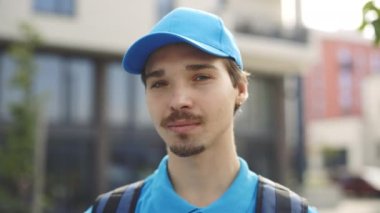 Sırt çantalı ve şapkalı mavi üniformalı beyaz erkek kurye. Yakışıklı mutlu postacı kameraya bakıyor. Erkek Sevkiyatının Yakın Görüntüsü