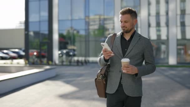 年轻的商人穿着正式的黑色西装走在写字楼中心附近 一边用手机 一边回答工作中的问题 男老板在他的工作场所附近发短信 在智能手机咖啡上翻阅应用程序 — 图库视频影像