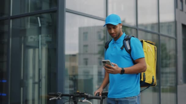 穿着制服站在商务中心附近骑自行车 使用智能手机和耳机 滚动客户订单的非洲裔美国人的肖像 食物派递 — 图库视频影像