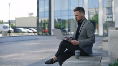 Genç iş adamı bankta oturuyor ve kulaklık takarak dizüstü bilgisayarla çalışıyor. Ofis merkezine yakın bir yerde çalışıyor. Çevrimiçi Yakışıklı Patron. Adam dışarıda otururken bilgisayarını kullanıyor.