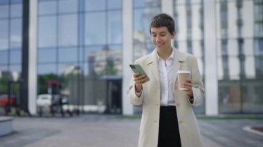 Gülümseyen İş Kadını Resmi Kahverengi Takım Elbisesiyle Yürüyor, Cihazı Kullanıyor ve İş Konuşmalarına Yanıt Veriyor. Kadın Patron, Smartphone Holding Coffee 'deki Uygulamalarla Mesajlaşıyor ve Kaydırıyor