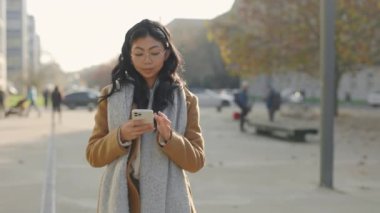 Neşeli, Asyalı genç bir bayan dışarıda yürüyor. Akıllı telefonunu tutuyor ve sarılıyor. Sosyal medyada Gadget Kullanarak Mesaja Cevap Veren Genç Kadın. Halk ve Teknoloji Konsepti