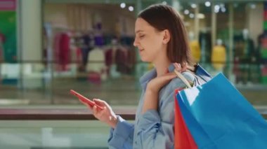 Alışveriş kolik kadın, alışveriş merkezinde yürürken telefonla konuşuyor. Renkli Kağıt Alışveriş Torbası ile Cep Telefonu Konuşan Kadın Alıcı. Alışveriş merkezine giden gülen kız. Yan Görünüm