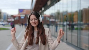 Neşeli Genç Kafkas Kadın Komik Dans ve Modern Şehir Binası 'nın arka planında Kulaklıkla Müzik Dinlerken ve Akıllı Telefon Cep Telefonu Tutarken. Açık havada.